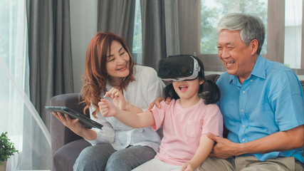 Asian grandparents and granddaughter using virtual reality and tablet play games at home. Senior Chinese, grandpa and grandma happy relax with young girl lying on sofa in living room concept.