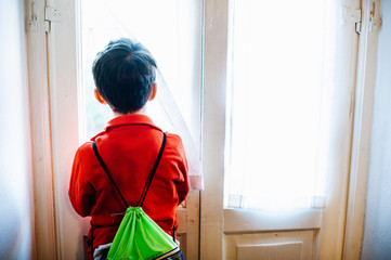 child with backpack looks out the window at home