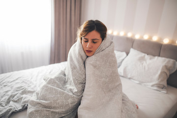 Sick girl sitting on the bed wrapped in blanket.