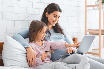 Fototapeta premium Pregnant mother and daughter shopping online, using laptop