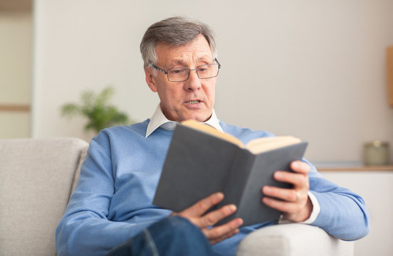 Retired Man Reading Book Sitting On Couch At Home