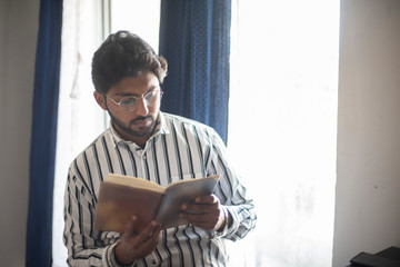 Young and handsome Indian brunette man in striped formal shirt and glasses reading a book while...