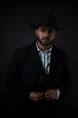 Fashion portrait of an young and handsome Indian Bengali brunette man with striped formal shirt, black suit and cowboy hat in black copy space background. Indian lifestyle and fashion photography.