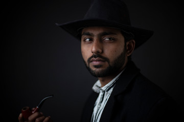 Fashion portrait of an young and handsome Indian Bengali brunette man with striped formal shirt, black suit, hat and a pipe in black copy space background. Indian lifestyle and fashion photography.