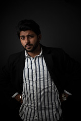 Fashion portrait of young and handsome Indian Bengali brunette man with striped formal shirt, black suit in black copy space background. Indian lifestyle and fashion photograph