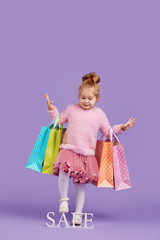 Portrait of a happy little child girl on a purple background holding shopping bags, package. Looking at the camera. Sale concept