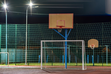 Outdoors mini football and basketball court with ball gate and basket surrounded with high protective fence brightly illuminated with spotlight lamps at night.