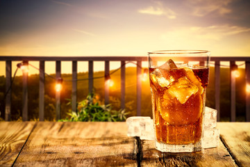 Cold ice tea in glass.Free space for your decoration on wooden table cover of ice cubes.Summer orange sunset time and blurred background of balcony. 