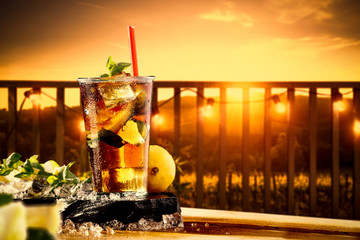 Cold ice tea in glass.Free space for your decoration on wooden table cover of ice cubes.Summer orange sunset time and blurred background of balcony. 