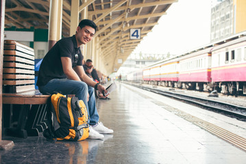 Cheap Travel and Lifestyle concept.Holiday Time,Young Solo Traveler man wearing sneaker and sitting at train station.Asian Backpacker waiting train alone and plan trip in summer time with smart phone