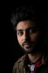 Obraz premium Fashion portrait of young and handsome Indian Bengali brunette man with casual camouflage shirt showing facial expression in black copy space background. Indian lifestyle and fashion photography