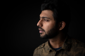 Fashion portrait of young and handsome Indian Bengali brunette man with casual camouflage shirt showing facial expression in black copy space background. Indian lifestyle and fashion photography