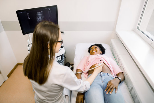 Young Caucasian lady doctor performing examination of abdomen with ultrasonography device for her young African female patient