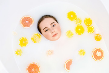 Obraz na płótnie Canvas Pretty girl in bath with milk and orange, lemon, grapefruit slices. Top view, only face under water. Natural make up.