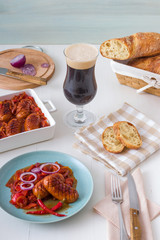 The plate with portion of baked sausages in dark beer with red onion, red and yellow paprika, red hot chilli, twisted rosemary ciabatta bread and glass of dark beer.