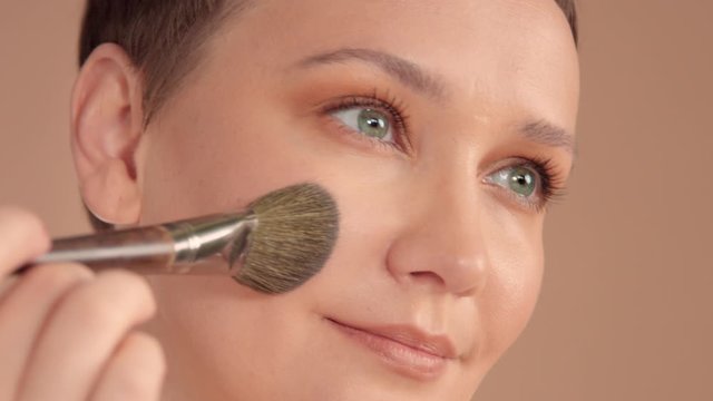 Caucasian woman with short haircut and natural beige makeup in studio on beige background put a makeup facial powder using a makeup brush. Watching a side. closeup