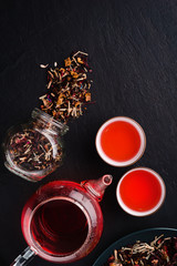 Fototapeta na wymiar Aromatic fresh brew natural red fruit tea in two bowls ,glass teapot,glass jar and plate with dry tea leaves on a black concrete background