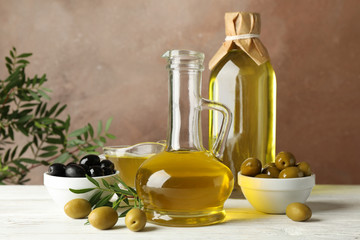 Bottle and jug with olive oil and olives against brown background, close up