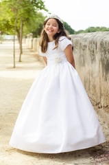 Fototapeta na wymiar Little girl in communion dress smiles with her arms crossed behind. Full body photography of the dress, sunbath