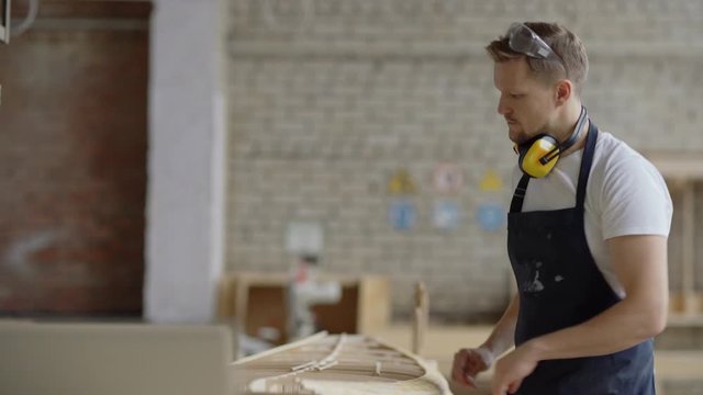 Middle aged professional carpenter constructing wooden boat model and using laptop computer to check plan drawing in carpentry shop