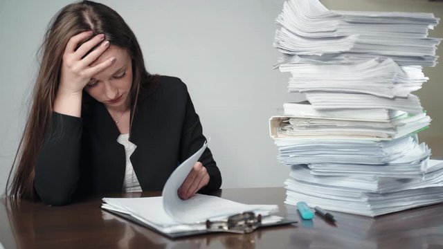 A pile of unfinished documents and files laying on desk in the office. Stressed and tired female office accountant sitting in front of paperworks and holding her head with hands, nervous worker having