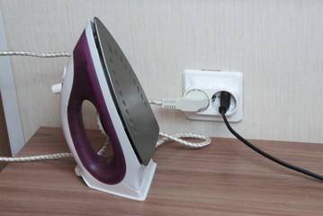 A lilac electric iron stands on a table plugged into a wall outlet