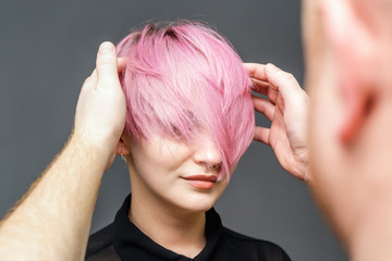 Fototapeta premium Hairdresser is checking short pink hairstyle of young woman on gray background, close up.