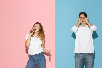 She's singing, he's shocked. Young and happy man and woman in casual clothes on pink, blue bicolored background. Concept of human emotions, facial expession, relations, ad. Beautiful caucasian couple.