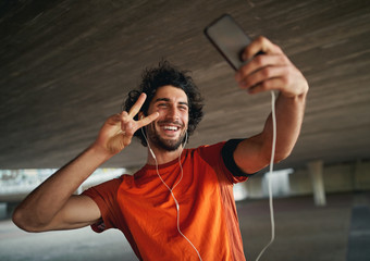 Portrait of a smiling young and handsome man with mobile smartphone pulling a peace sign and taking...
