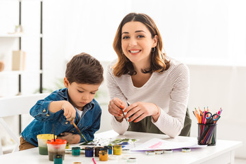 happy woman looking at camera while drawing with paints together with son