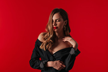 Fototapeta premium Attractive stylish woman with red lips and wavy hair wearing black dress with bare shoulders posing with closed eyes and smile over red background