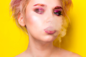The blonde girl exhales smoke and looks into the frame on a yellow background.