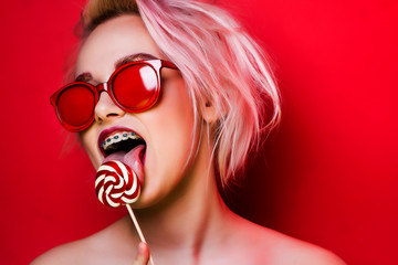 Girl with pink hair and braces licks a candy on a red background. Photo of a blonde in red glasses with candy. - obrazy, fototapety, plakaty