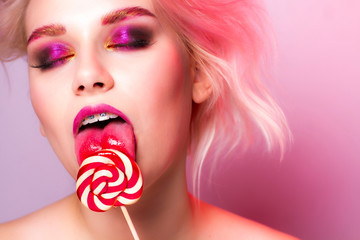 A model with bright makeup licks a candy on a pink background with eyes closed.