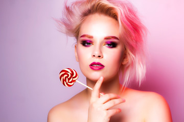A blonde with bright makeup on a pink background looks into the frame and holds a candy in her hands.