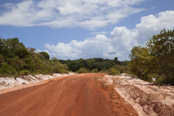A dirt road made of red clay with sides of white sand on a clear Sunny day against a background of blue sky and clouds. Landscape, jungle, savanna, world tourism. 