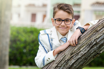 Close up portrait of a brunette little boy leaning on a tree trunk wearing a fashion white sailor...