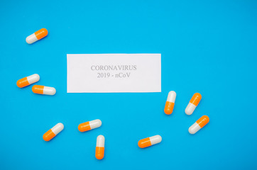 white sheet of paper with the inscription Coronavirus among orange-and-white capsules on a blue background. Medical background, top view