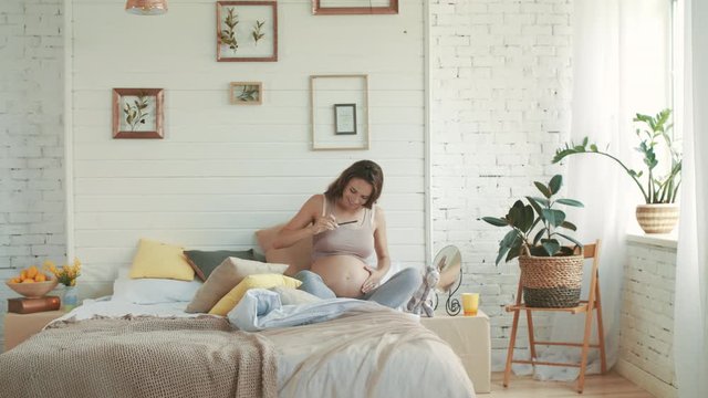 Young pregnant woman taking belly selfie at home. Happy belly mother sitting bed