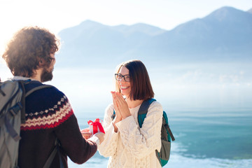 Young man proposing to woman and giving gift box. Happy couple of travelers at winter sea beach by mountains. Happy students celebrate engagement outdoor. Romantic marriage proposal. Lifestyle moment.