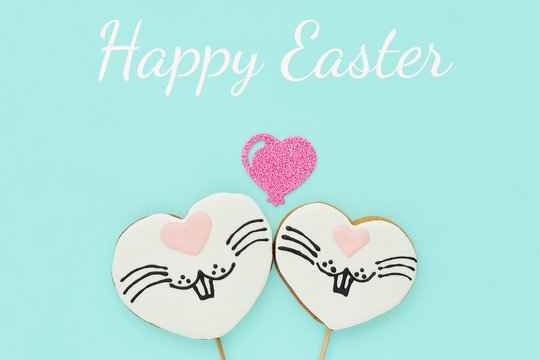 Greeting card Happy Easter, two gingerbread cookies on a stick in the shape of a heart with a drawn rabbit face and a shiny heart on a blue background...