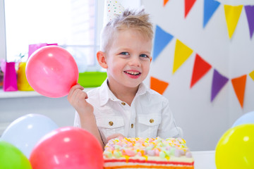 Portrait of blonde caucasian boy smiling at camera near birthday rainbow cake. Festive colorful background with balloons.