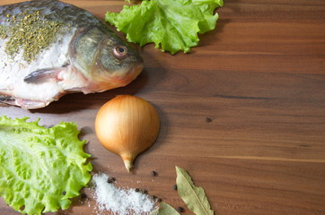 On the table lies a fresh, gutted river fish, crucian carp, onions, lettuce, salt, spices.