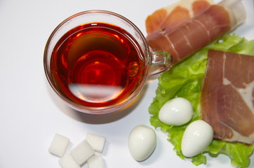 Morning. Healthy breakfast. On a white background is a glass cup with black tea, salad leaves, boiled quail eggs, jamon.