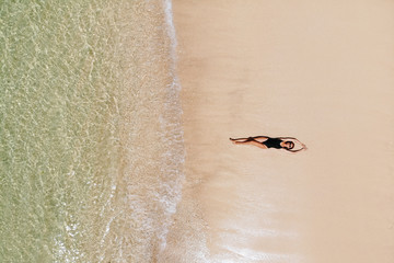 Aerial view of young woman in black bikini lying on beach with white sand, foaming waves of the Indian Ocean. Bali Island, Indonesia. Photo from drone. Space for text.