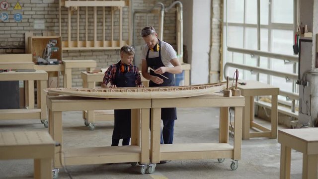 Wide shot of middle aged professional woodworker explaining to little son how to make wooden boat model standing at workbench in carpentry shop