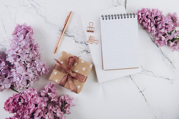 Spring flat lay top view home office workspace - gift and notebook with lilac flowers on a marble desk background