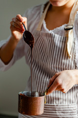 Beautiful young girl in beige apron is preparing delicious organic hot chocolate in old vintage ladle. Soft daylight, lifestyle photography, process of tasty drink creation. Close up