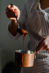 Beautiful young girl in beige apron is preparing delicious organic hot chocolate in old vintage ladle. Soft daylight, lifestyle photography, process of tasty drink creation. Close up