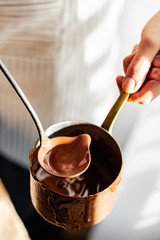 Beautiful young girl in beige apron is preparing delicious organic hot chocolate in old vintage ladle. Soft daylight, lifestyle photography, process of tasty drink creation. Close up